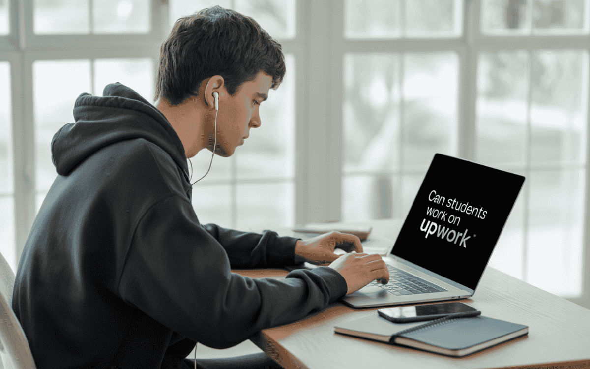 Student wearing hoodie typing on laptop with "Can students work on Upwork?" displayed on screen in sunlit room.